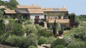 Una casa de dos pisos con tejado de baldosas, rodeada de vegetación exuberante y terreno rocoso.