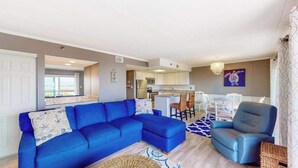 Un salon avec un canapé bleu, un fauteuil bleu et un coin repas avec des chaises blanches.