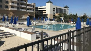 Un espace piscine avec des chaises longues et des parasols, un parking souterrain et un immeuble à plusieurs étages doté de balcons.