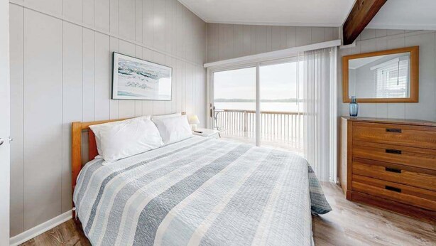 Une chambre à coucher comprenant un lit, une commode, une grande fenêtre avec des rideaux, un tableau encadré accroché au mur et une poutre en bois au plafond.