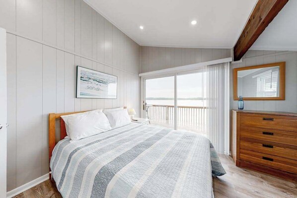 A bedroom with a bed, a dresser, a large window with curtains, a framed picture on the wall, and a wooden ceiling beam. - 14 Clipper on the Bay (Ocean Pines)