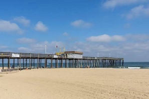 A beach with a wooden pier extending into the ocean, a small building at the end, and a clear sky.