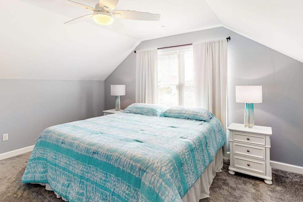 Un dormitorio con cama, dos mesitas de noche, ventilador de techo y ventana con cortinas.