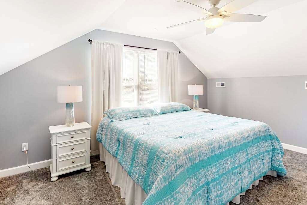 Quarto com cama, duas mesas de cabeceira, ventilador de teto e janela com cortinas.