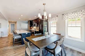 A modern kitchen with dark cabinetry, a central island, and a dining area with a wooden table and chairs. - New home steps from Northside Park with shared pools, tennis & pickleball (Ocean City)