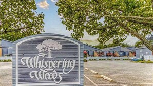 A sign for 'Whispering Woods' with a tree design, surrounded by a parking lot and residential buildings. - Ground-floor condo with pool & fenced patio - 4 blocks to the beach (Ocean City)