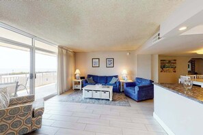 A living room with a sofa, coffee table, and lamps; a balcony with a view; and a kitchen area. - Fountainhead Towers 310 (Ocean City)