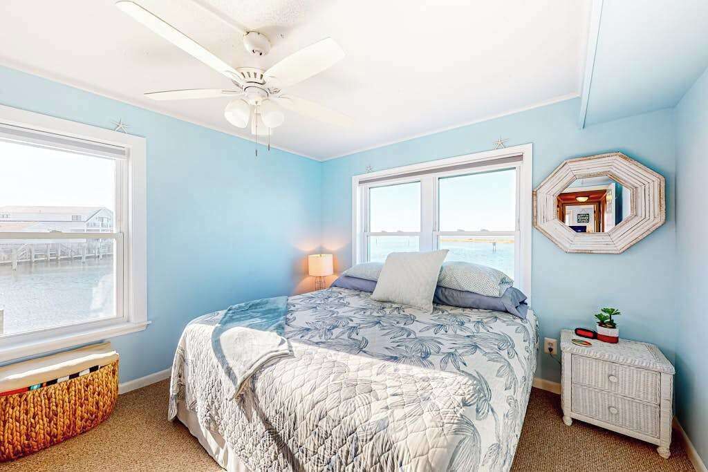 A bedroom with a bed, two windows, a ceiling fan, a nightstand, and a wall-mounted mirror.