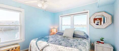 A bedroom with a bed, two windows, a ceiling fan, a nightstand, and a wall-mounted mirror.