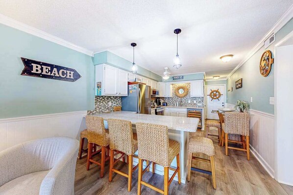 A kitchen with a central island, white cabinets, wicker chairs, and a 'BEACH' sign on the wall. - Bayspot A-101 (Ocean City)