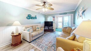 Sala de estar con dos sofás, un televisor, un ventilador de techo y una pared con un letrero que dice 'Beach'.