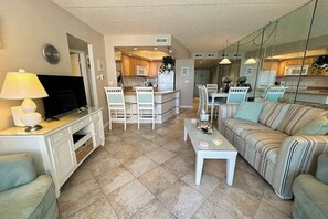 A living room with a TV, sofa, coffee table, and a kitchen area with a dining table and chairs. - Spacious oceanfront condo with indoor pool, sauna, game room, great beach access (Ocean City)