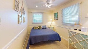 A bedroom with a bed, ceiling fan, two windows with blinds, a bedside table with a lamp, and wall-mounted picture frames.
