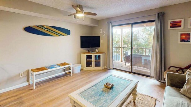 Een woonkamer met een plafondventilator, een televisie op een kast, een bank, een bijzettafeltje en een schuifdeur naar een balkon.
