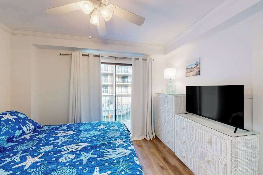 Une chambre à coucher avec un lit, un ventilateur de plafond, une télévision, une fenêtre avec des rideaux et une vue sur un bâtiment à l’extérieur.
