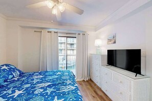 Ein Schlafzimmer mit Bett, Deckenventilator, Fernseher, einem Fenster mit Vorhängen und Blick auf ein Gebäude.