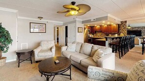 Un salon avec un canapé, des fauteuils, une table basse, un ventilateur de plafond et, au fond, un coin cuisine.