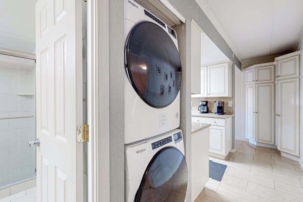 Une buanderie équipée d’un lave-linge et d’un sèche-linge à chargement frontal, un coin cuisine avec des placards et une douche visible à travers une porte ouverte.
