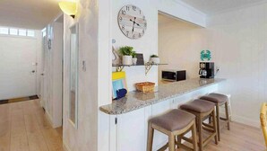 A kitchen with a clock, bar stools, a coffee machine, and a small shelf with decorative items. - Camelot 603B (Ocean City)