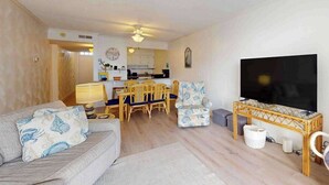A living room with a sofa, a television, a dining area with chairs, and a kitchen in the background. - Camelot 603B (Ocean City)