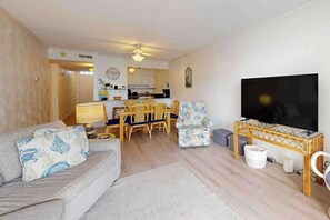 A living room with a sofa, a television, a dining area with chairs, and a kitchen in the background. - Luxury Ocean View Condo w/Shared Olympic Pool, Tennis, balcony & WiFi (Ocean City)