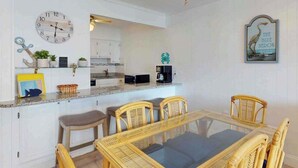 A modern kitchen with white cabinets, a dining table with chairs, a clock, and a coffee machine. - Camelot 603B (Ocean City)
