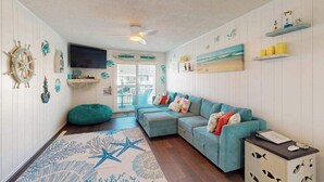 A living room with a teal sofa, wooden flooring, nautical decor, a flat-screen TV, and a ceiling fan. - Sunny canal side condo - w/balcony, short walk to the beach, boardwalk, & parks (Ocean City)