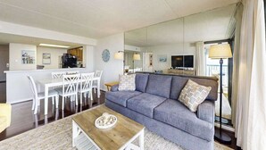 En stue med sofa, sofabord, spiseområde med stole, et fjernsyn og et køkken.