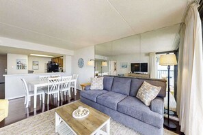 A living room with a sofa, a coffee table, a dining area with chairs, a TV, and a kitchen area. - Charming Beach Condo w/Balcony, beach & bay views, Pools, Gym, Tennis, W/D & A/C (Ocean City)