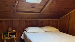 Une chambre avec un puits de lumière, des murs en bois, un lit avec une literie blanche et une petite table de chevet avec des livres et une plante.