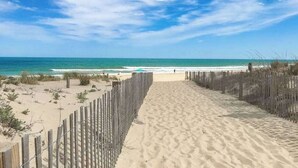 Ein sandiger Pfad am Strand, der zum Meer führt, mit einem Holzzaun an einer Seite und einem strahlend blauen Himmel darüber.