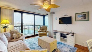 Ein Wohnzimmer mit Sofa, Sessel, Hocker, Deckenventilator, Fernseher und einem Balkon mit Blick.