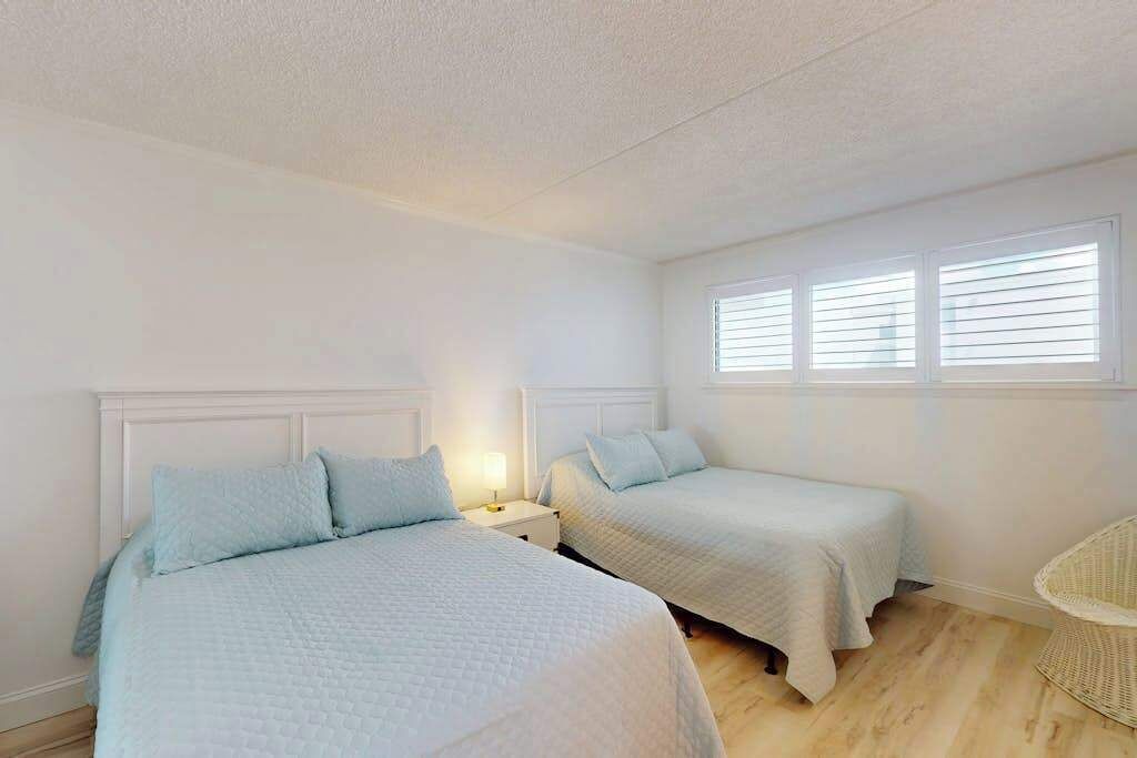A bedroom with two beds, a window with blinds, a wicker chair, and a nightstand with a lamp.