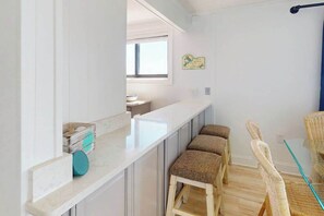 A kitchen with a white countertop, wooden stools, wicker chairs, and a window. - Camelot 501B (Ocean City)