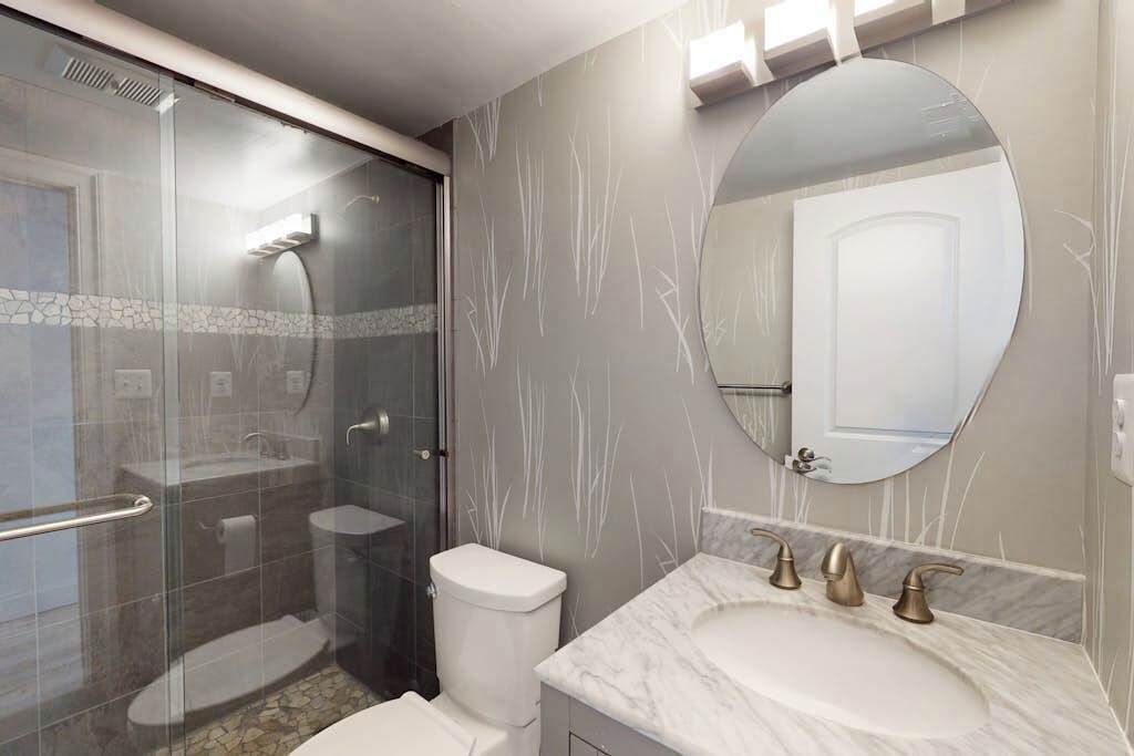 A bathroom with a glass shower enclosure, a white toilet, a marble sink with three faucets, and a large mirror.