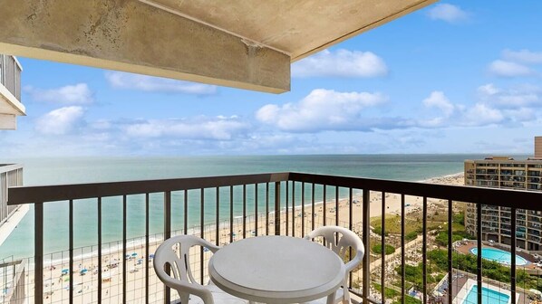 A balcony with a table and chairs, overlooking a beach and a pool. - Serene oceanfront condo with beach views, pools, gym, tennis & sauna (Ocean City)