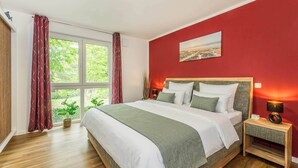 Ein Schlafzimmer mit einem großen Bett, einem Nachttisch mit Lampe, einem Fenster mit Vorhängen und einem Bild an der Wand.