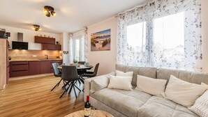 Ein modernes Wohnzimmer mit Sofa, Esstisch mit Stühlen, Küchenzeile mit Schränken und einem großen Fenster mit Vorhängen.