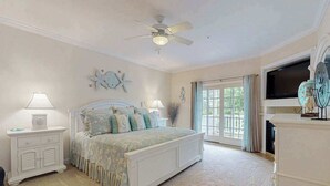 Une chambre à coucher comprenant un lit, des tables de chevet, un ventilateur de plafond, une grande fenêtre et une cheminée.