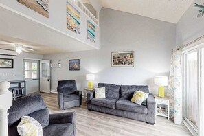 Un salon avec un canapé gris, deux fauteuils, une petite table et une lampe. Les murs sont ornés de tableaux encadrés et il y a une fenêtre avec des rideaux.