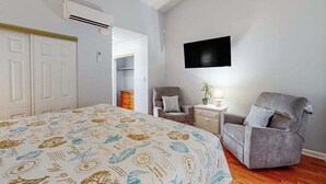 Quarto com cama, poltrona cinza, TV na parede e ar condicionado.