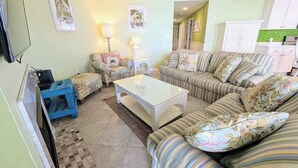 A living room with a sofa, a coffee table, a blue chair, and a TV. - Spacious - Family-Friendly Beach Condo w/Balcony, A/C, Shared Rooftop Pool & Gym (Ocean City)