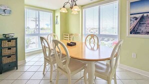 A dining area with a round table, white chairs, and a view through large windows. - Spacious - Family-Friendly Beach Condo w/Balcony, A/C, Shared Rooftop Pool & Gym (Ocean City)