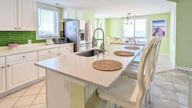 A kitchen with white cabinets, a stainless steel refrigerator, a sink, and a dining area with a table and chairs. - Spacious - Family-Friendly Beach Condo w/Balcony, A/C, Shared Rooftop Pool & Gym (Ocean City)