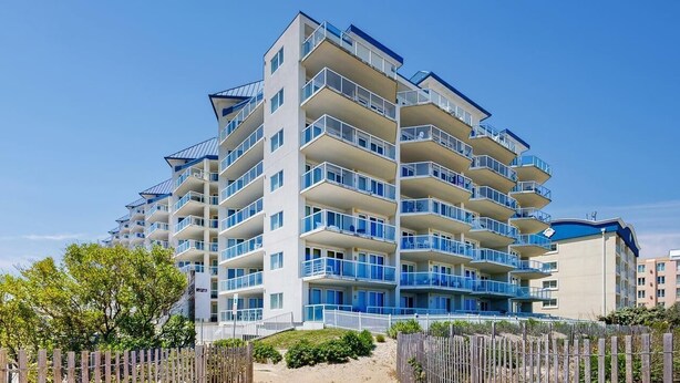 Un hotel de varios pisos con balcones, una playa de arena frente al establecimiento y un cielo azul despejado.
