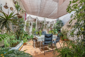 En overdækket terrasse med et bord og stole, omgivet af forskellige potteplanter.
