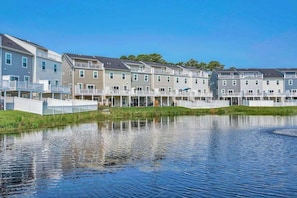 A row of modern multi-story buildings with balconies, a calm water body in the foreground, and a clear blue sky.