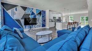 A modern living room with a large blue sofa, a flat-screen TV, and a geometric wall mural. - Sea Oaks Village 12442 #5 (Berlin)