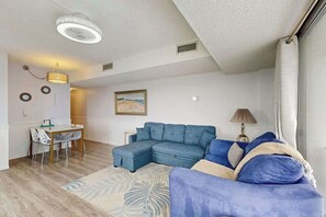 Ein Wohnzimmer mit Ecksofa, Esstisch mit Stühlen, einer Lampe und einem Bild an der Wand.