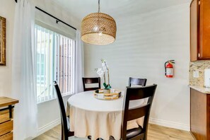 A dining area with a round table, chairs, a hanging light fixture, a window with blinds, and a fire extinguisher on the wall. - Oceanside Escape with AC / Walk to Beach, Coffee & Dining (Oceanside)
